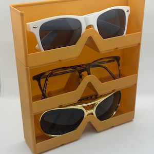 May include: An orange sunglasses organiser with three compartments, each holding a pair of sunglasses. The sunglasses include white-framed, tortoiseshell, and gold-framed styles. The organiser is made of a solid, opaque material.