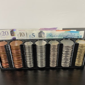 May include: A black plastic coin holder with six compartments, each filled with a stack of British coins. The holder has a slot at the top for inserting notes, and the notes are visible in the image. The notes are a £20, a £10, and a £5 note.