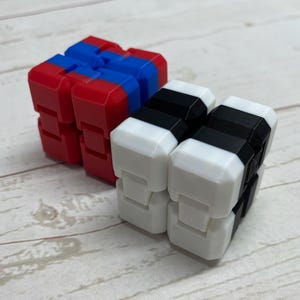 May include: Two fidget cubes, one red, blue and black, and the other black and white. The cubes are made of plastic and have a textured surface.