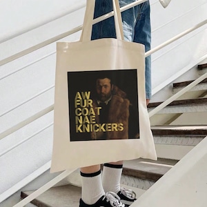 May include: A cream tote bag with a black and gold graphic. The graphic shows a portrait of a man in a fur coat with the words "AW FUR COAT NAE KNICKERS" in gold lettering. The bag is held by a person on a staircase.