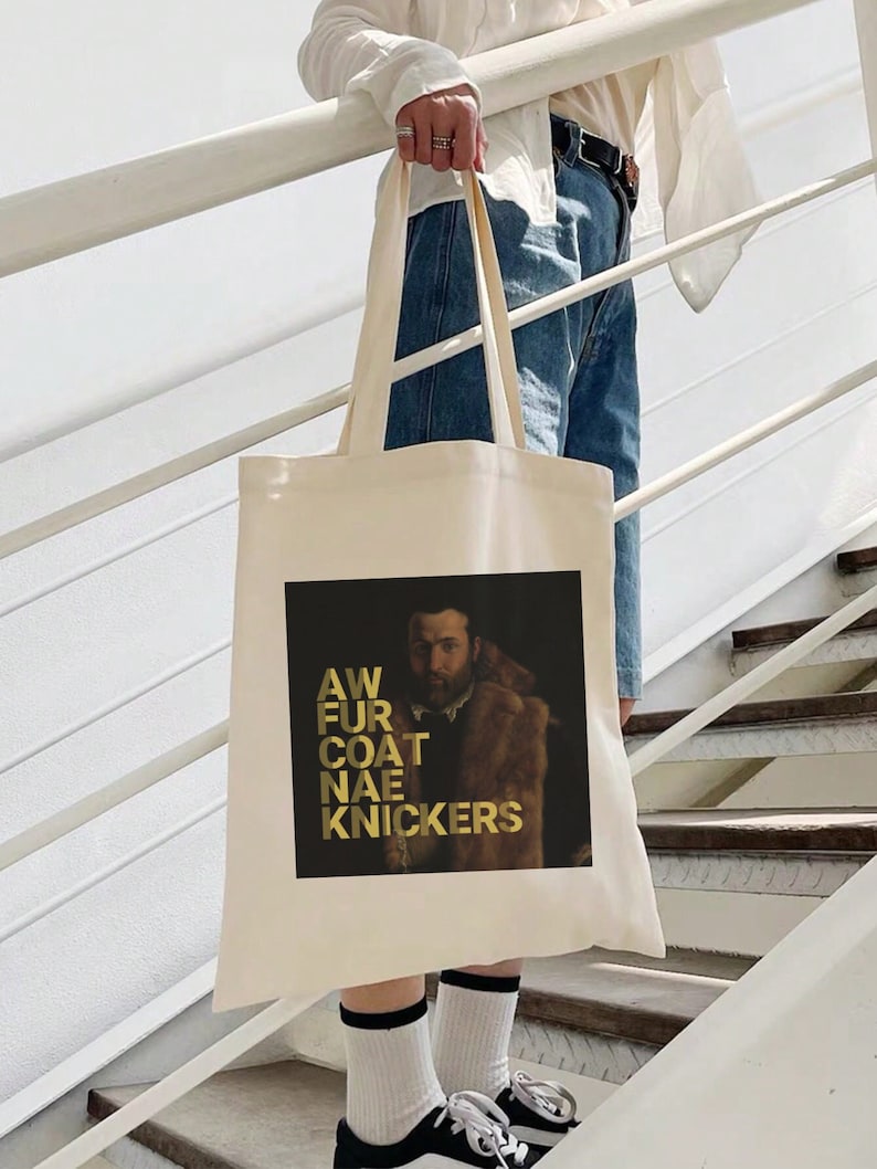 May include: A cream tote bag with a black and gold graphic. The graphic shows a portrait of a man in a fur coat with the words "AW FUR COAT NAE KNICKERS" in gold lettering. The bag is held by a person on a staircase.