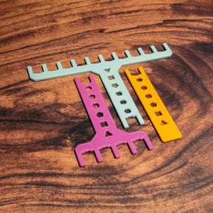 Puede incluir: Tres herramientas de tejido de plástico con mangos de diferentes colores: azul claro, rosa y amarillo. Cada herramienta tiene un extremo en forma de peine y una fila de agujeros a lo largo del mango.