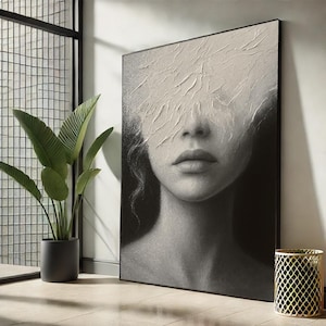 May include: Monochrome portrait print of a woman's face, with textured white paint obscuring the eyes. The artwork is in a black frame, leaning against a white wall. A potted plant and a gold metal container are in the background.