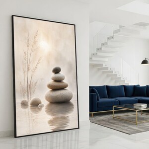 Op de afbeelding: Een ingelijste kunstdruk met een serene scène van gestapelde stenen in een rustige omgeving. Het kunstwerk toont gladde, afgeronde stenen die op elkaar in evenwicht zijn, met zacht, diffuus licht en waterreflecties. Het kleurenpalet is neutraal, met beige- en grijstinten.