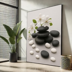 May include: A framed art print featuring a zen-inspired composition. Black and white stones are arranged with white blossoms and green leaves against a light gray background. The artwork is displayed in a room with a potted plant and a decorative gold-colored metal container.