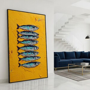 May include: A framed artwork featuring a textured yellow background with seven stylized fish painted in shades of blue, gray, and white. The artwork is displayed in a modern living room setting.