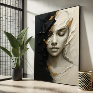 May include: A framed abstract artwork depicting a woman's face with closed eyes. The piece is split vertically, with a textured black side and a white side. Gold accents highlight the transition. The artwork is displayed near a potted plant.