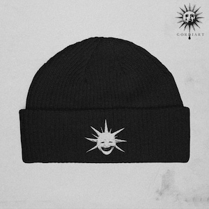 May include: Black knit beanie featuring a white smiling sun graphic on the front. The beanie has a ribbed texture and a folded brim. The brand name "GORDIART" is printed in white on the upper right corner.