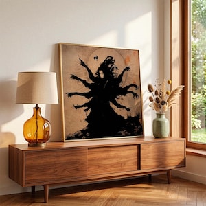May include: A framed art print featuring a black silhouette of a multi-armed figure with the Japanese character for "God". The artwork is set against a beige background with a golden frame. The print is displayed on a wooden cabinet with a lamp and vase.