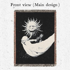 Puede incluir: Manta negra con una ilustraci&oacute;n detallada de una figura solar con una larga lengua, sosteniendo una figura en un pa&ntilde;o blanco. El texto "Front view (Main design)" est&aacute; en la parte superior. La manta tiene un borde con flecos.