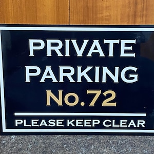 May include: A rectangular black sign with a white border featuring the white text PRIVATE PARKING, the gold text No.72, and the white text PLEASE KEEP CLEAR at the bottom, separated by a horizontal white line.