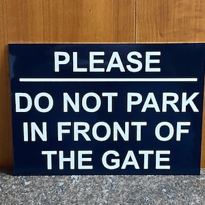 May include: A navy blue rectangular sign with white lettering. The sign states "PLEASE DO NOT PARK IN FRONT OF THE GATE". The sign is set against a wooden background and is designed to be a clear warning.