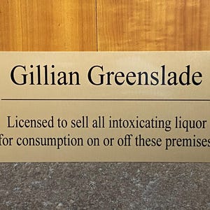 May include: A rectangular gold-coloured metal sign with black engraved text that reads: Gillian Greenslade, Licensed to sell all intoxicating liquor for consumption on or off these premises. The sign features a horizontal line separating the name.