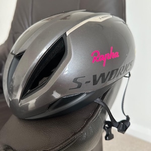 May include: A gray Specialized S-Works cycling helmet with the pink Rapha logo. The helmet has a sleek, aerodynamic design with black accents and a chin strap. The helmet is designed for road cycling.