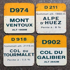 May include: Four vintage-style road signs with yellow and white rectangular designs. Each sign displays a route number, mountain name, and altitude in meters. The signs are for Mont Ventoux, Alpe d'Huez, Col du Tourmalet, and Col du Galibier.