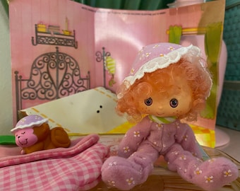 May include: A pink and white doll wearing pink and white floral print pyjamas and a pink and white floral print hat. The doll has pink hair and blue eyes. The doll is sitting on a pink and white checked blanket.