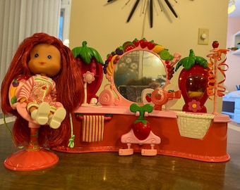 May include: A pink and red strawberry-themed vanity set with a mirror, a hairdryer, a stool, and a doll with red hair. The vanity has a pink and white striped towel hanging on it.