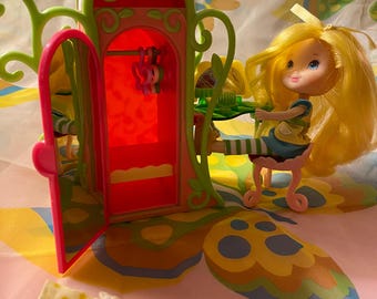 May include: A yellow doll with long blonde hair is sitting on a pink and green stool in front of a red and green toy wardrobe. The wardrobe has a mirror on the door and a hanger inside. The doll is holding a green brush. There is a yellow and white dress on the floor in front of the wardrobe.