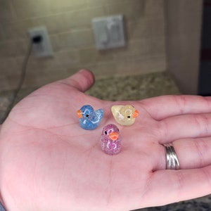 May include: Three small glass duck figurines. The ducks are blue, yellow, and pink with glitter accents.