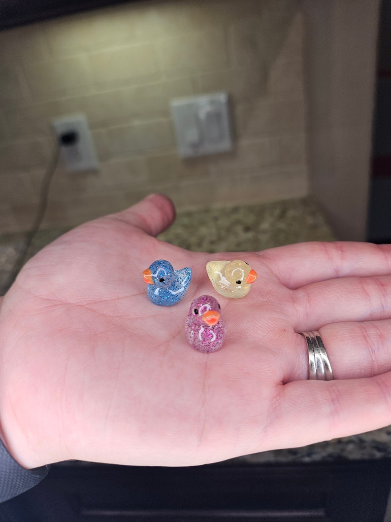 May include: Three small glass duck figurines. The ducks are blue, yellow, and pink with glitter accents.