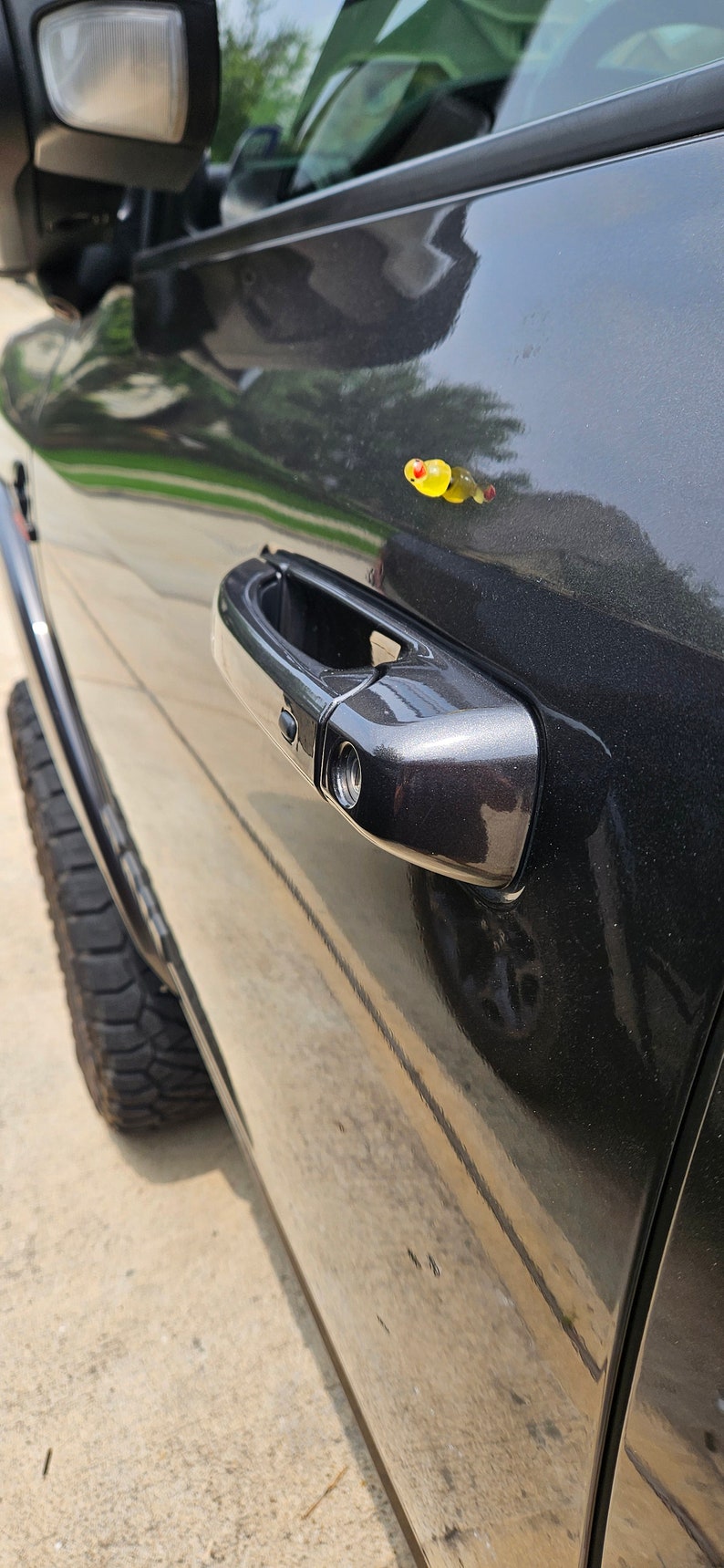 May include: A black truck door with a chrome door handle and a small yellow object on the door.