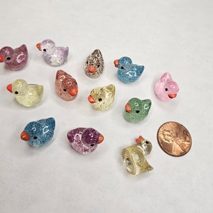 May include: A collection of 12 small, glittery, duck-shaped figurines in various colours, including blue, pink, green, yellow, and purple. The figurines are arranged on a white surface.