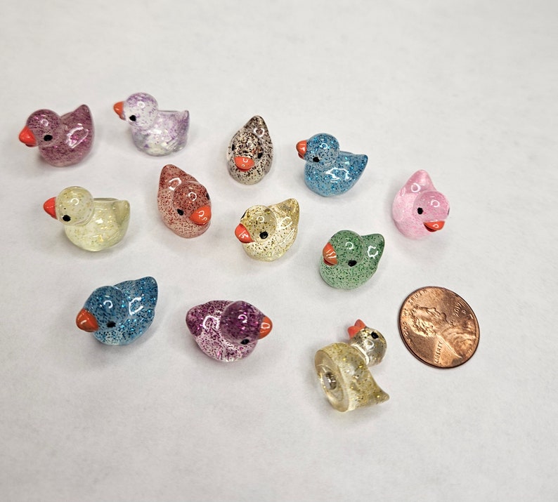 May include: A collection of 12 small, glittery, duck-shaped figurines in various colours, including blue, pink, green, yellow, and purple. The figurines are arranged on a white surface.
