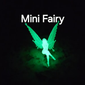 May include: A glowing green fairy figurine with detailed wings and a flowing dress, set against a black background. The text "Mini Fairy" is displayed in white above the fairy. The fairy appears to be illuminated from within.