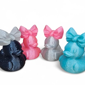 May include: Five decorative duck figurines in a range of colours. The ducks are black, pink, silver, and blue, each with a matching bow on their heads. The figurines are made of a textured material.