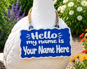 May include: A white goose statue wearing a blue rectangular sign with a scalloped white border. The sign features white text that reads Hello my name is Your Name Here and is suspended from the goose's neck by a braided tan rope.
