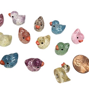 May include: A collection of 12 small, glittery duck figurines in various colours, including pink, blue, green, purple, and gold. The ducks are about the size of a UK penny.