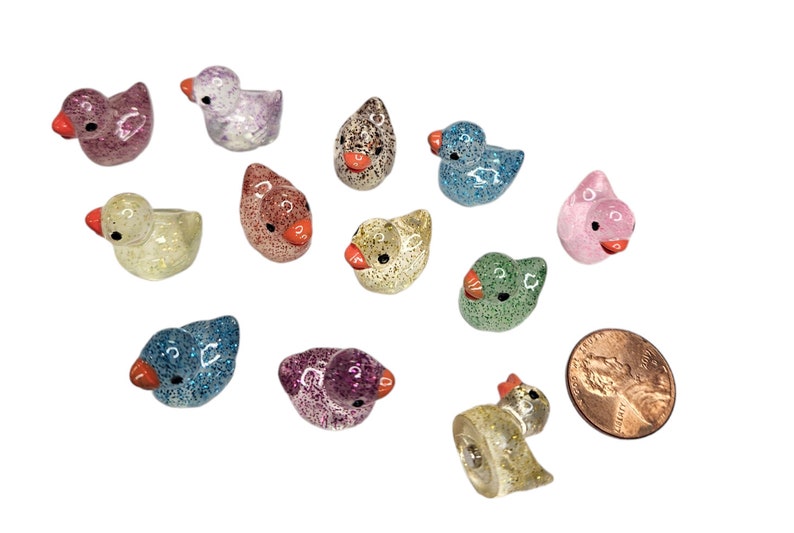May include: A collection of 12 small, glittery duck figurines in various colours, including pink, blue, green, purple, and gold. The ducks are about the size of a UK penny.