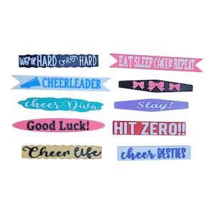 May include: A selection of colourful cheerleading-themed ribbons, each bearing a different phrase. The ribbons are in shades of pink, blue, black, purple, and gold, with phrases such as "Work Hard, Cheer Hard," and "Cheerleader."