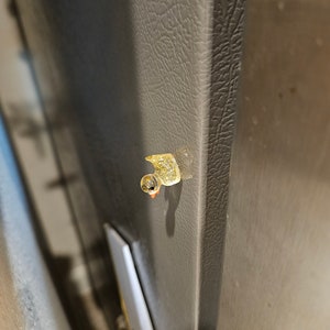 May include: A small, yellow rubber duck with a black eye and orange beak is attached to a grey refrigerator door.