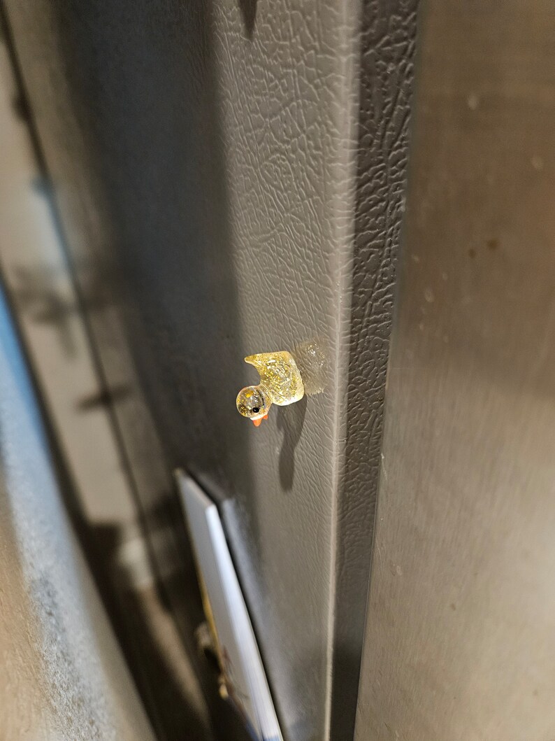 May include: A small, yellow rubber duck with a black eye and orange beak is attached to a grey refrigerator door.