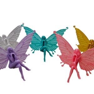 Könnte beinhalten: Fünf Feenfiguren mit Flügeln, jede in einer anderen Farbe: lila, weiß, türkis, rosa und gold. Die Feen sitzen mit ausgebreiteten Flügeln.