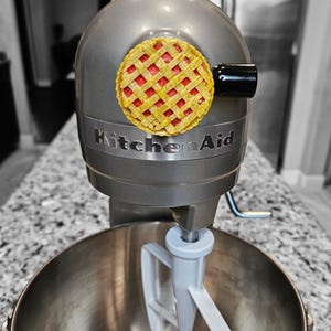 Puede incluir: Una batidora de pie KitchenAid plateada con una pegatina de masa de pastel roja y amarilla en el lateral. La batidora tiene un tazón blanco y un accesorio de paleta blanco.