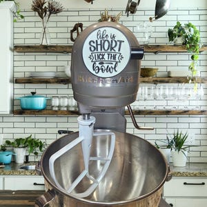 Op de afbeelding: Een zilveren KitchenAid-standmixer met een witte kom en een witte paddle-hulpstuk. De mixer heeft een ronde sticker aan de zijkant met de tekst "life is short lick the bowl".