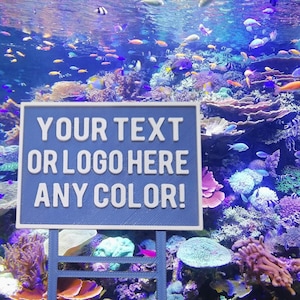 May include: A blue sign with white text that reads "YOUR TEXT OR LOGO HERE ANY COLOR!" The sign is in front of a colorful coral reef with many fish swimming around.