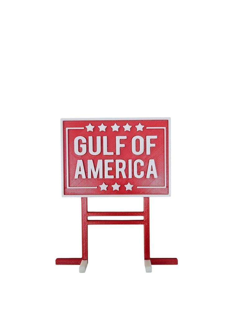 K&ouml;nnte beinhalten: Ein rotes Schild mit wei&szlig;em Text "GULF OF AMERICA" und einem wei&szlig;en Rand. Das Schild hat einen roten St&auml;nder mit wei&szlig;en F&uuml;&szlig;en. Das Schild hat f&uuml;nf Sterne &uuml;ber dem Text und drei darunter.