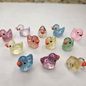 May include: A collection of 12 small, glittery duck figurines in various colours, including pink, blue, green, yellow, and purple. The ducks have orange beaks and are made of glass or plastic.