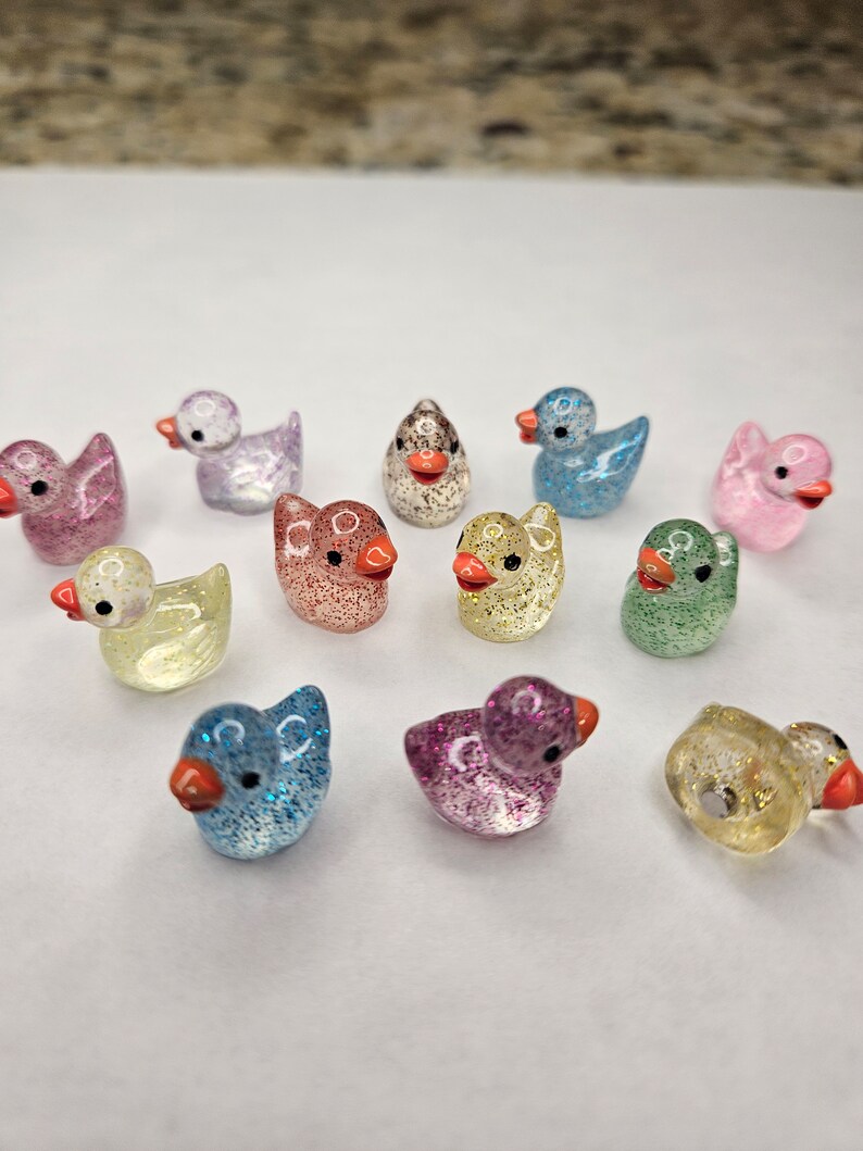 May include: A collection of 12 small, glittery duck figurines in various colours, including pink, blue, green, yellow, and purple. The ducks have orange beaks and are made of glass or plastic.