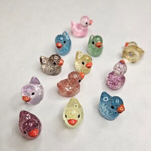 May include: A collection of 12 small, glittery rubber duck figurines in various colours, including pink, blue, green, yellow, and purple. The ducks are all facing forward and have a small orange beak.