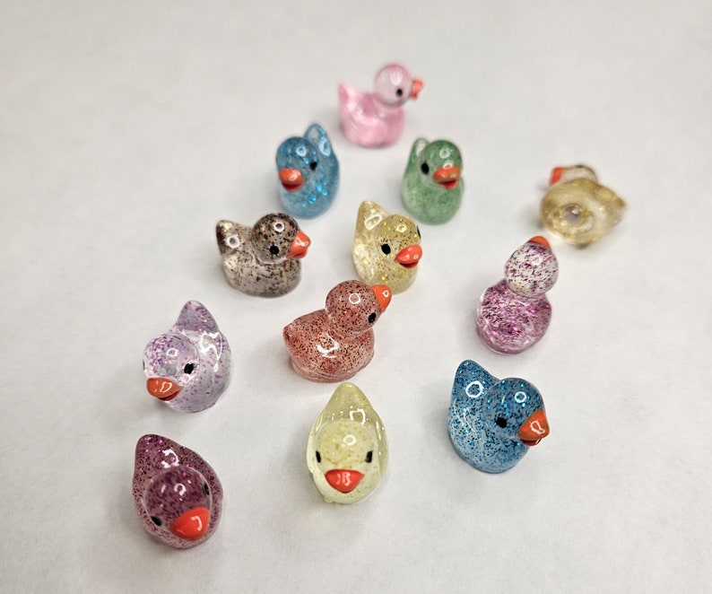 May include: A collection of 12 small, glittery rubber duck figurines in various colours, including pink, blue, green, yellow, and purple. The ducks are all facing forward and have a small orange beak.