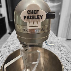 Puede incluir: Una batidora de pie KitchenAid plateada con un gancho de masa blanco. Una pegatina blanca con el texto "CHEF PAISLEY" está pegada a la batidora.