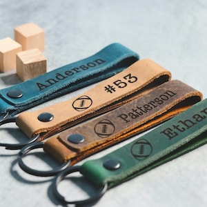 May include: Four leather keychains with a football design and personalized names. The keychains are in blue, brown, dark brown, and green.