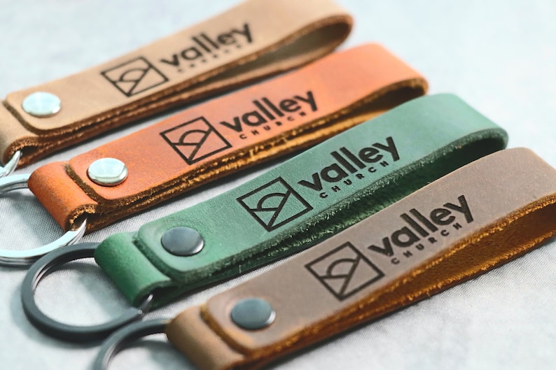 May include: Four leather keychains with a black metal ring. Each keychain has a different color: brown, orange, green, and tan. The keychains are embossed with the text 
