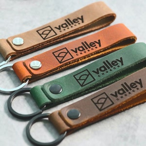 May include: Four leather keychains with metal key rings. Each keychain has a brown leather strap with the text "valley church" and a graphic of a mountain range inside a square. The keychains are in different colors: brown, orange, green, and dark brown.