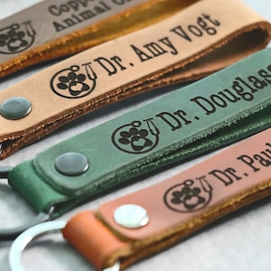 May include: Close-up of several leather keychains in various colors, including brown, tan, and green. Each keychain features engraved text with a veterinarian theme, including a paw print and the title "Dr."