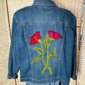 May include: A denim jacket with a floral design on the back. The flowers are painted in shades of red and pink, with green stems and leaves. The jacket is a medium wash blue and has a classic button-up design.